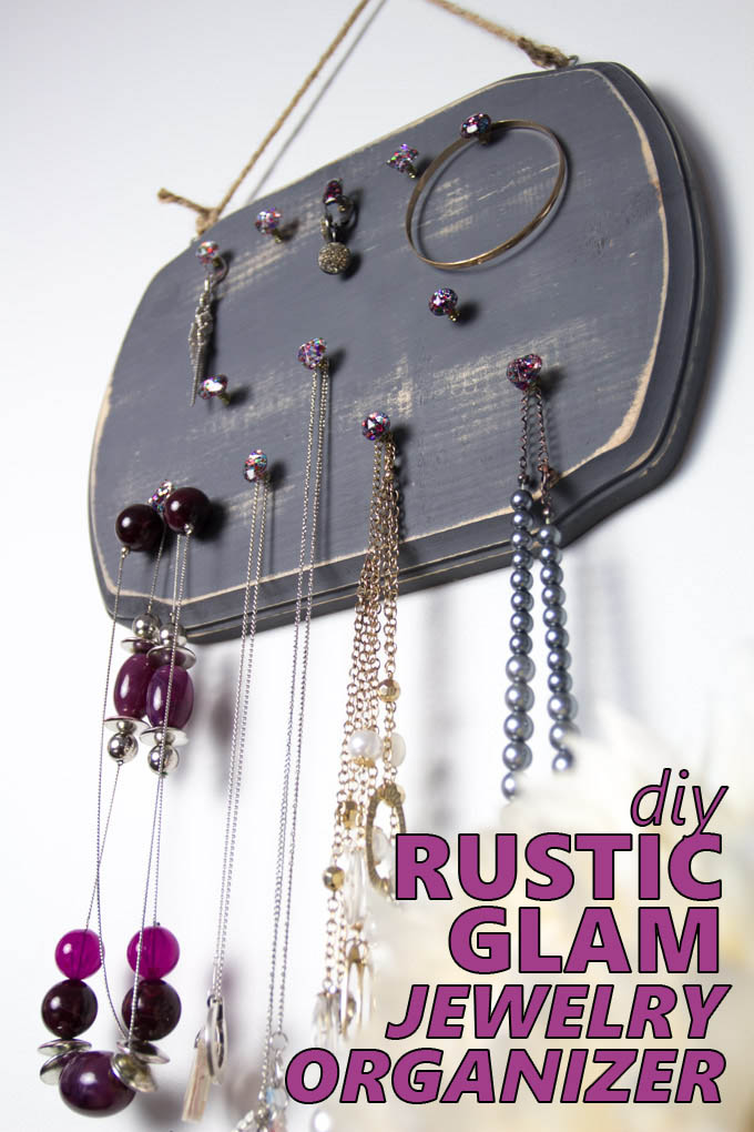 A distressed gray wood jewelry organizer hanging on a wall with jewelry