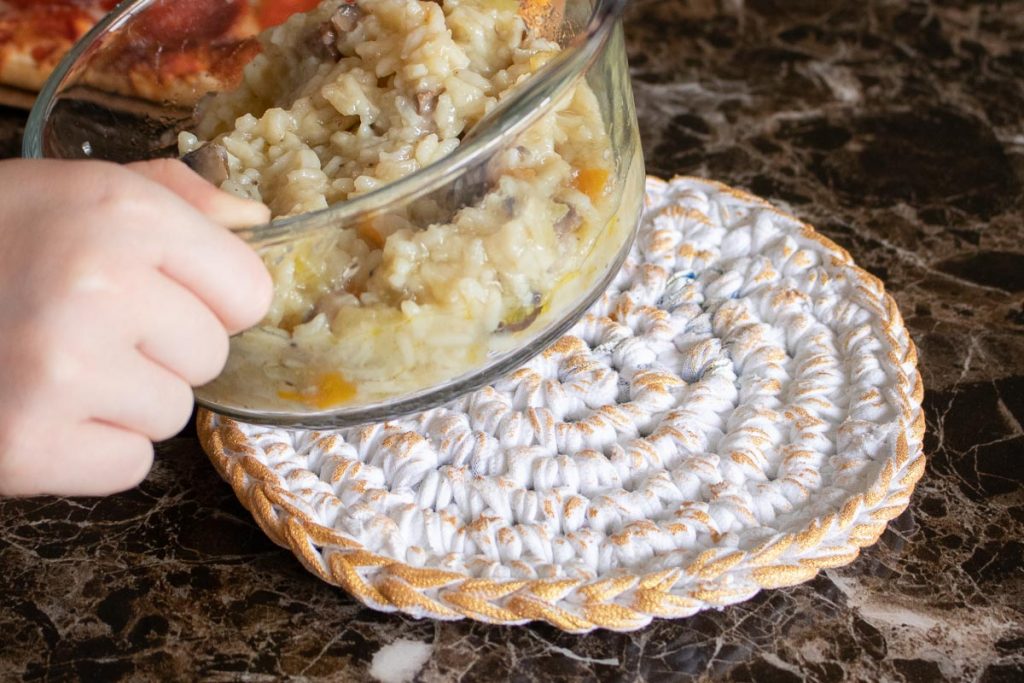 A clear bowl of food being placed onto a crochet heat pad painted with gold accents.